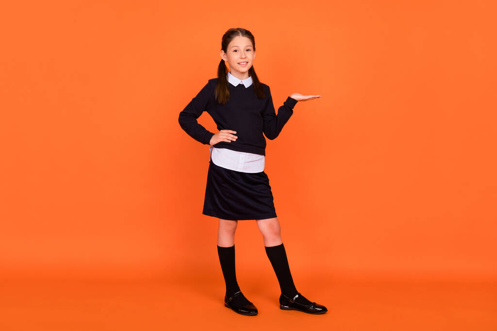 Fénykép imádnivaló iskolás kéz bemutató üres tér kopás egyenruha elszigetelt narancs színű háttér - Fotó, kép