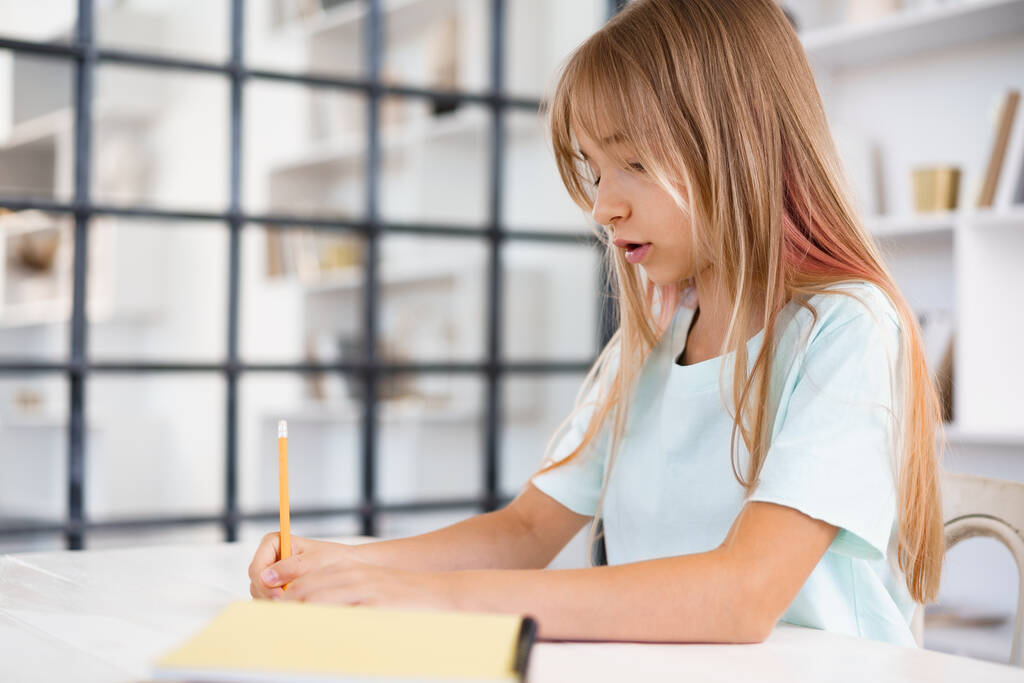 Девочка-подросток делает заметки в блокноте, делает домашнюю работу за столом дома - Фото, изображение