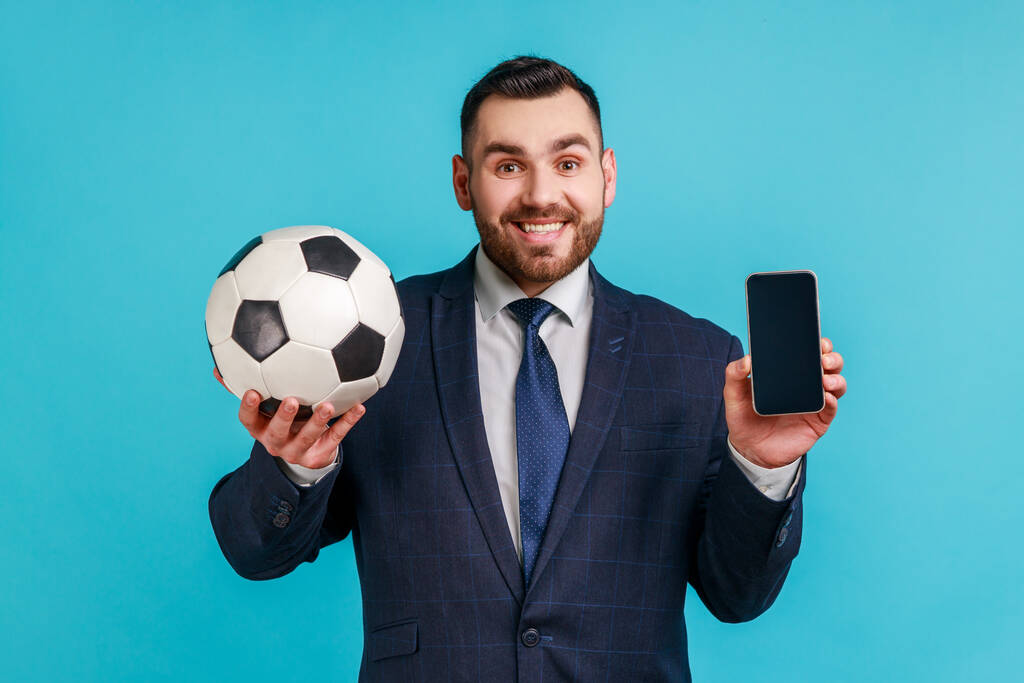 Hombre de negocios sonriente con traje de estilo oficial que sostiene el teléfono inteligente con el teléfono inteligente en blanco y la pelota de fútbol, apostando al fútbol. Estudio interior plano aislado sobre fondo azul. - Foto, Imagen