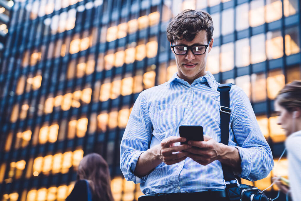 Bajo ángulo de trabajador masculino en camisa y gafas mensajes de texto en el teléfono de pie contra el alto edificio iluminado en Nueva York - Foto, Imagen