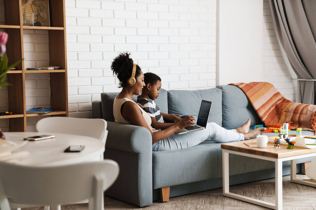 Negro feliz madre e hijo usando el ordenador portátil mientras está sentado en el sofá en casa - Foto, imagen