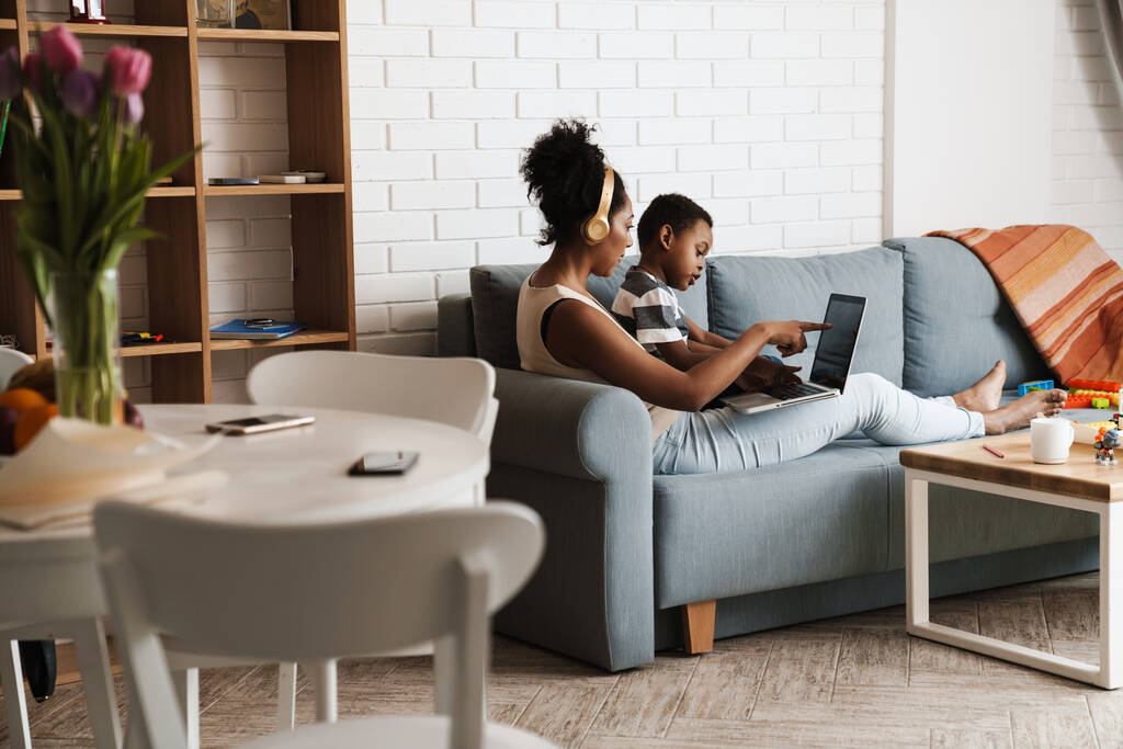 Чернокожие мать и сын пользуются ноутбуком, сидя дома на диване - Фото, изображение