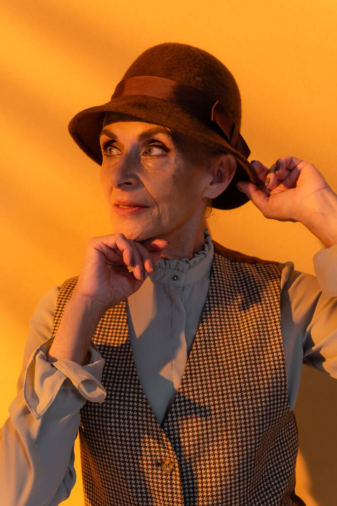 Portret van senior model in retro kleding en hoed op oranje achtergrond  - Foto, afbeelding