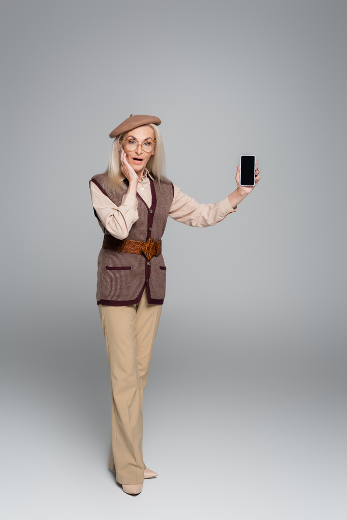 Úžasné starší žena drží smartphone s prázdnou obrazovkou na šedém pozadí  - Fotografie, Obrázek