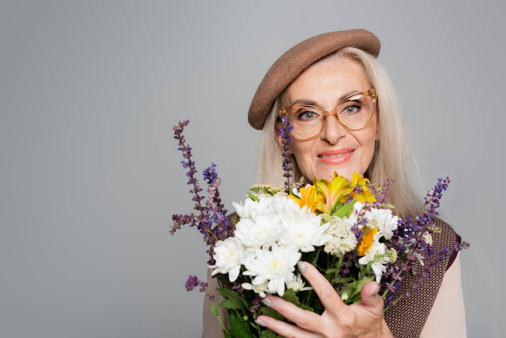 Smiling senior woman in beret and eyeglasses holding bouquet isolated on grey  - Photo, Image