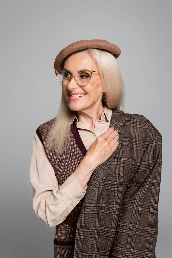Θετική και κομψή ηλικιωμένη γυναίκα κρατώντας σακάκι και κοιτάζοντας μακριά απομονώνονται σε γκρι  - Φωτογραφία, εικόνα