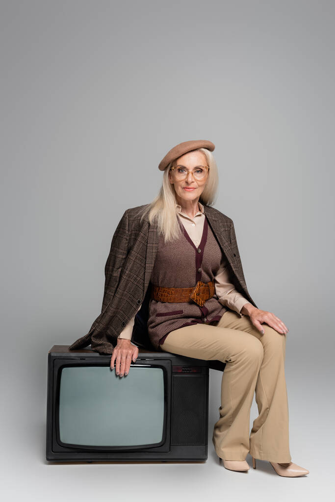 Mujer mayor en chaqueta a cuadros sentada en retro tv sobre fondo gris  - Foto, imagen