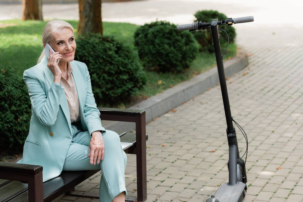 joyeuse femme d'affaires appelant sur le téléphone mobile sur le banc près de scooter électrique kick - Photo, image