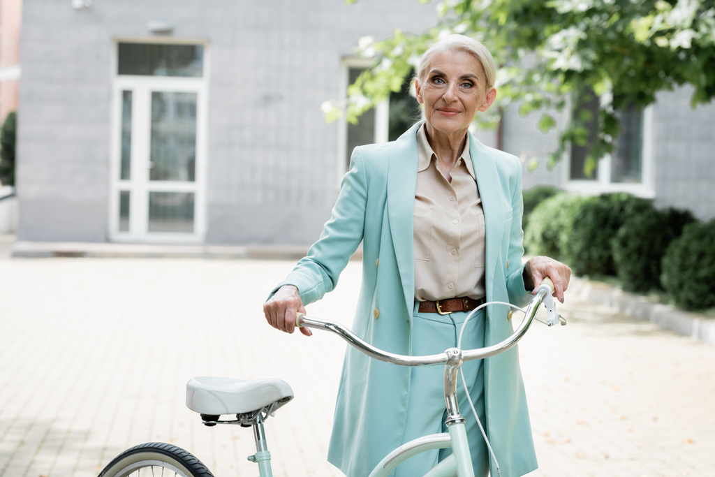 stijlvolle senior zakenvrouw glimlachend op camera in de buurt van fiets buiten - Foto, afbeelding