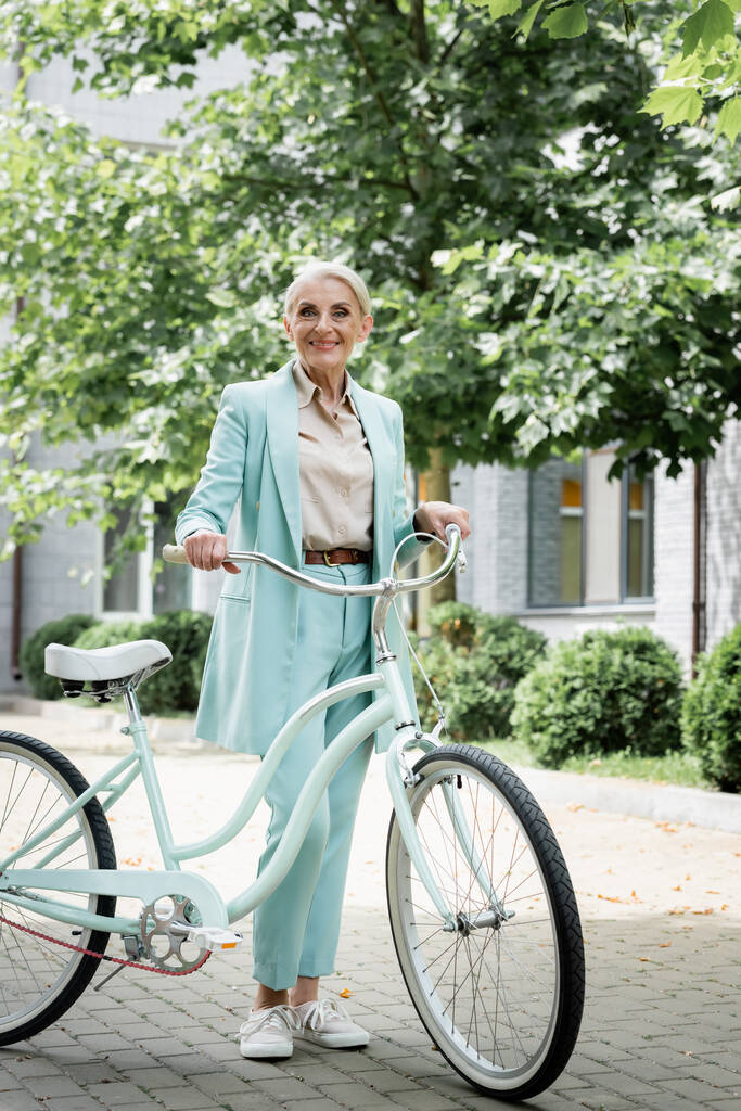 senior businesswoman in blue suit smiling near bicycle on urban street - Photo, Image