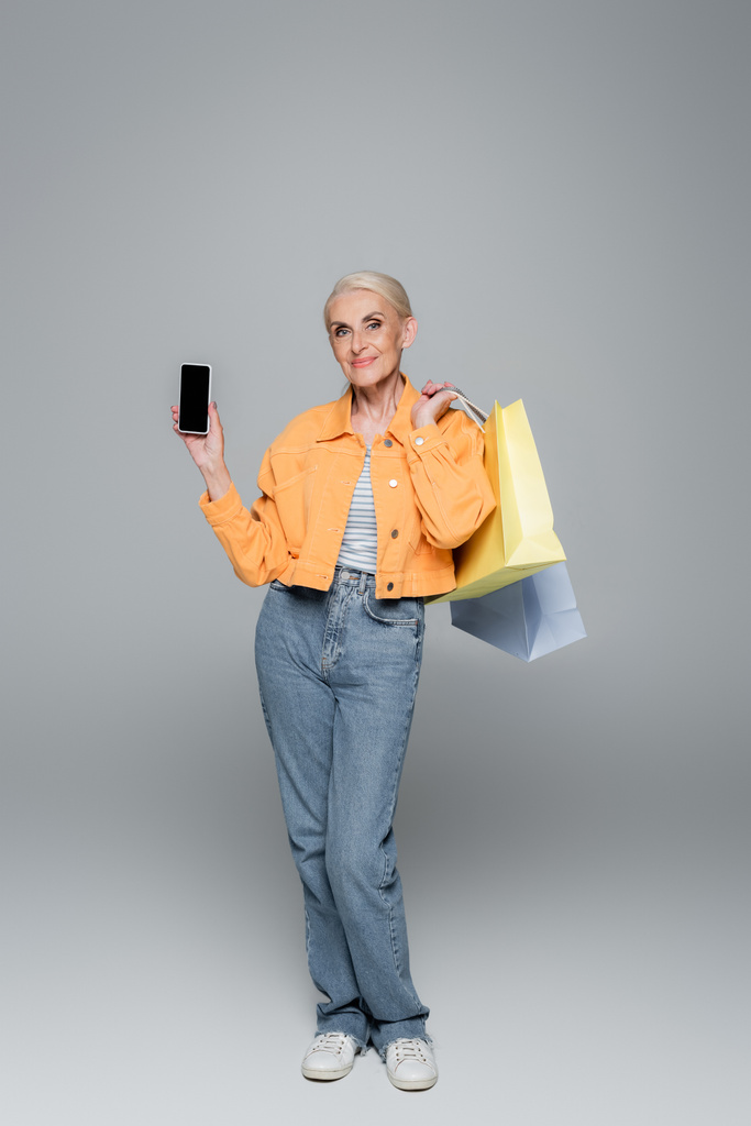 mujer anciana positiva posando con bolsas de compras y teléfono móvil en gris - Foto, Imagen
