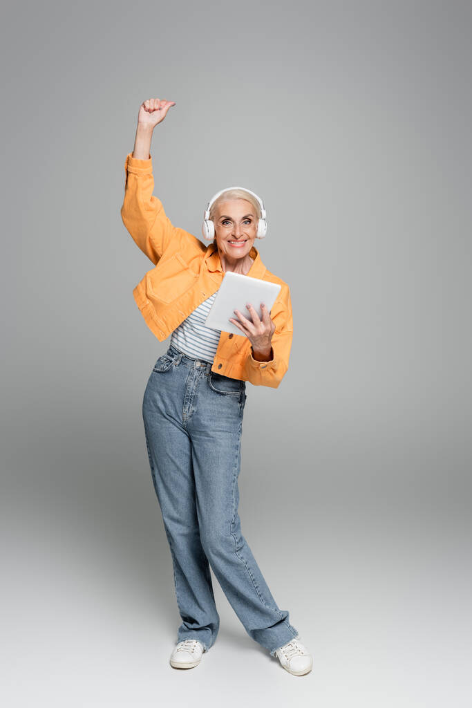 mulher idosa alegre em fones de ouvido segurando tablet digital e mostrando gesto de vitória em cinza - Foto, Imagem