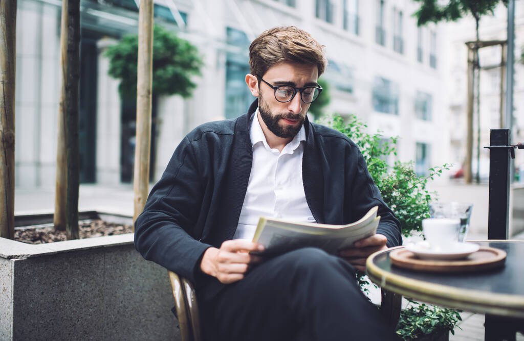 Nyugodt szakállas férfi sötét kabátban és világos ingben békésen olvas újságot, miközben az asztalnál ül egy csésze kávéval a szabadtéri kávézóban. - Fotó, kép