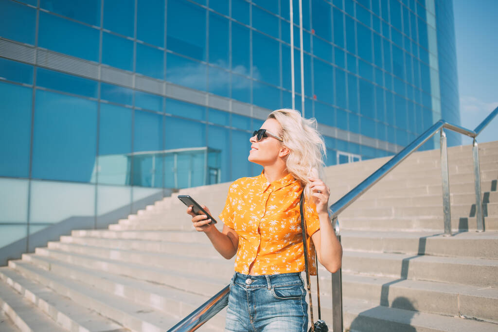 Беззаботная хипстерша в модных солнцезащитных очках, отводящая взгляд в ветреную погоду на улицу, 20-летняя блоггерша, держащая цифровые смартфоны в городской обстановке - Фото, изображение