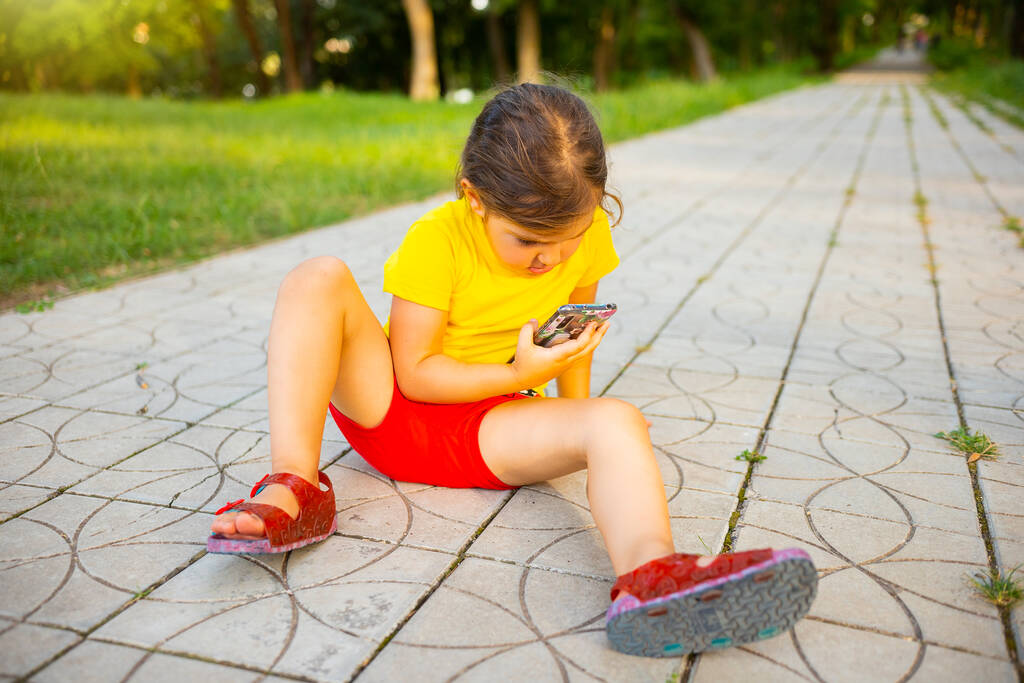 Holčička sedící na stezce v parku ve žlutém tričku a červených šortkách, držící smartphone v rukou a dívající se na něj. - Fotografie, Obrázek