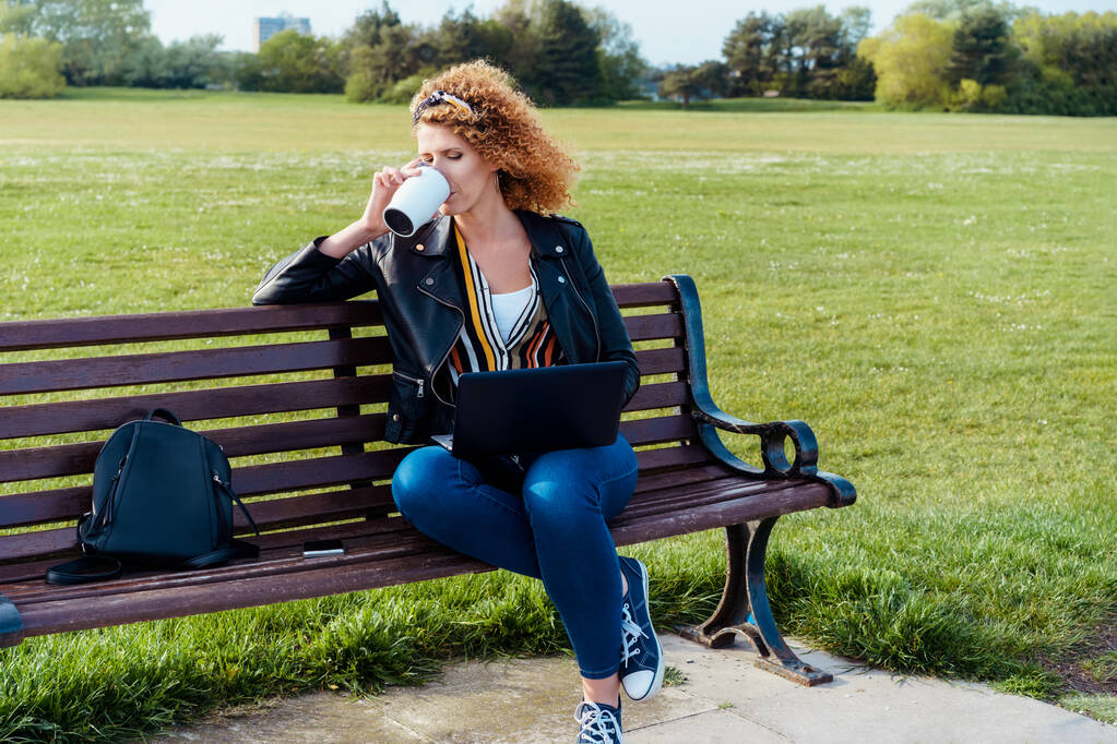 Una mujer vestida con ropa casual bebiendo café de una taza reutilizable y usando una computadora portátil al aire libre sentada en el banco en el parque público. Trabajo remoto, trabajo independiente. Redes. - Foto, Imagen