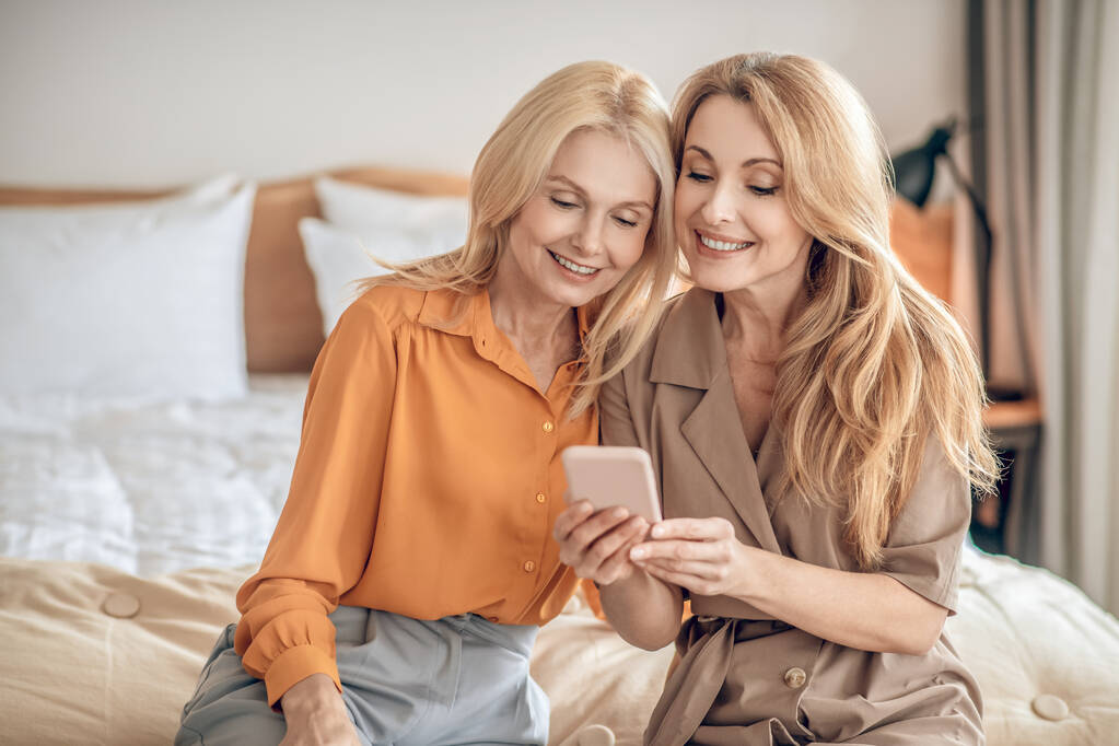 Zwei lächelnde blonde Frauen sitzen auf dem Bett und machen ein Selfie mit dem Smartphone - Foto, Bild