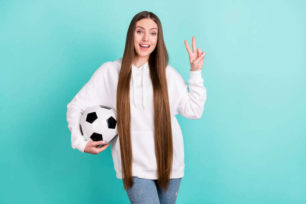 Фото позитивной молодой женщины держать за руку футбол сделать V-знак изолированы на пастельно-бирюзовом фоне - Фото, изображение