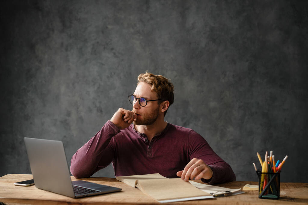 O homem pensativo sentado em uma mesa com papéis e um laptop enquanto trazia sua mão para o queixo no estúdio - Foto, Imagem