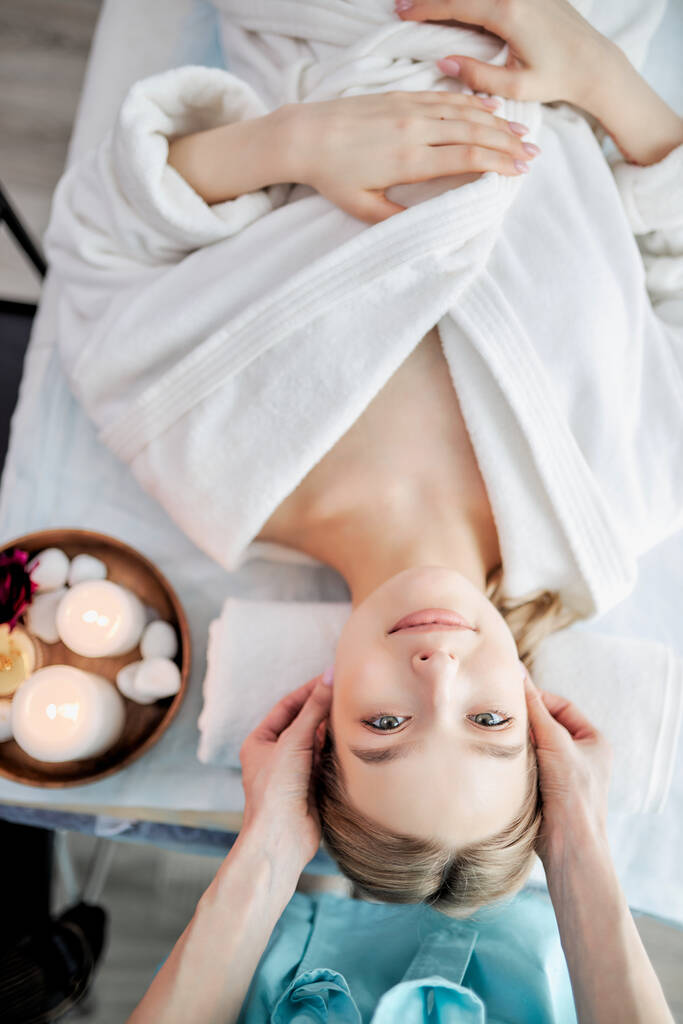 Вид зверху на позитивну кавказьку жінку, що розслабляється, а ретельний спеціаліст масажує її обличчя
 - Фото, зображення