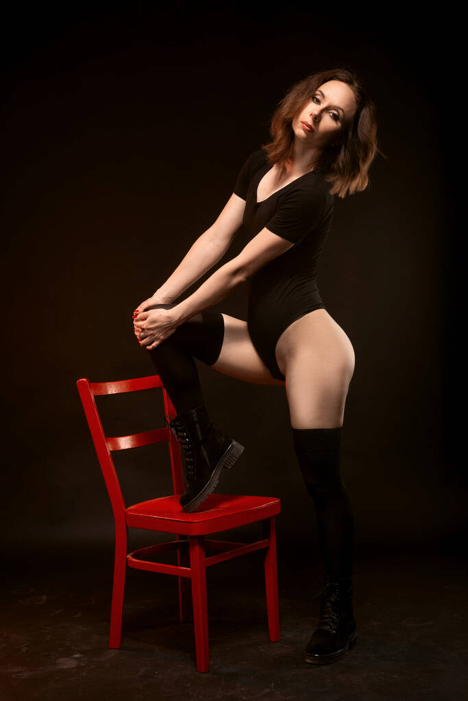 Szczupła kobieta w czarnym body i pończochach pozuje na czarnym tle w pobliżu czerwonego krzesła. sexy młoda kobieta. - Zdjęcie, obraz
