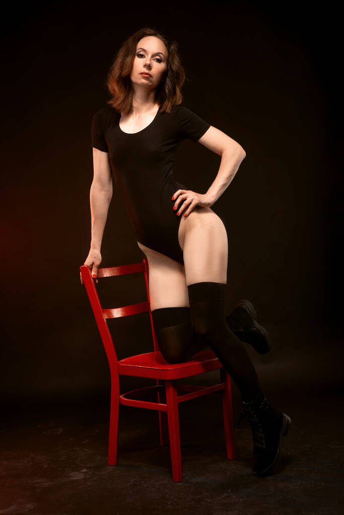 Szczupła kobieta w czarnym body i pończochach pozujących na czarnym tle na czerwonym krześle. sexy młoda kobieta. - Zdjęcie, obraz