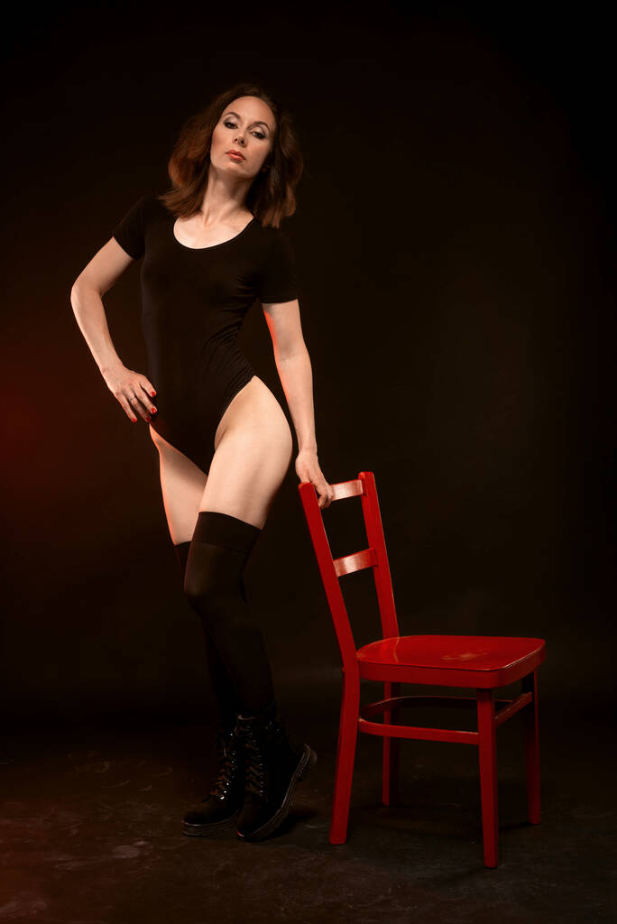 Una mujer delgada con un traje negro y medias posa sobre un fondo negro cerca de una silla roja. Sexy joven mujer. - Foto, imagen