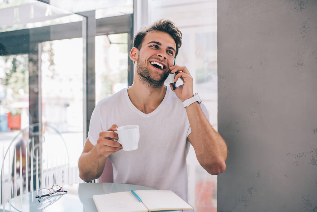 Onnellinen nuorekas komea mies valkoisessa t-paidassa, jolla on keskustelu älypuhelimella nauttien aamukahvista kahvilassa - Valokuva, kuva