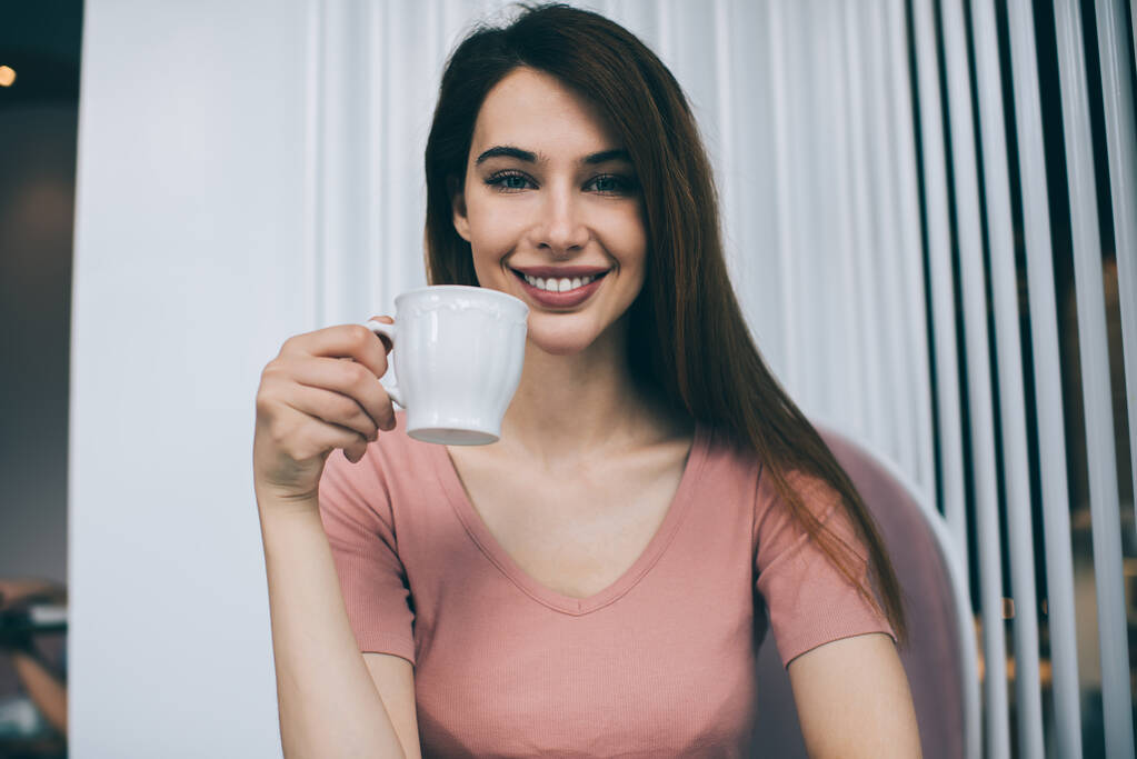 Morena bonita em camiseta rosa sorrindo alegremente e segurando pouca xícara branca enquanto olha para a câmera no fundo com persianas brancas - Foto, Imagem