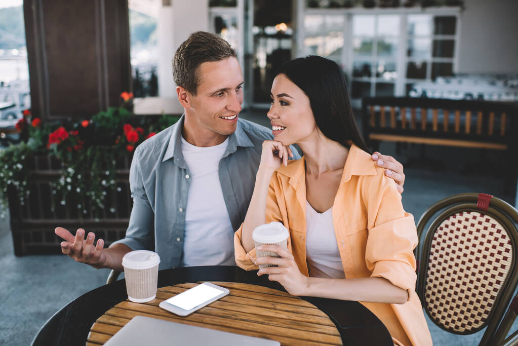 Happy laughing couple on date outside coffee shop with smartphone with empty screen man talking and resting hand on shoulder of agreeing girlfriend leaning on hand looking at each other - Photo, Image