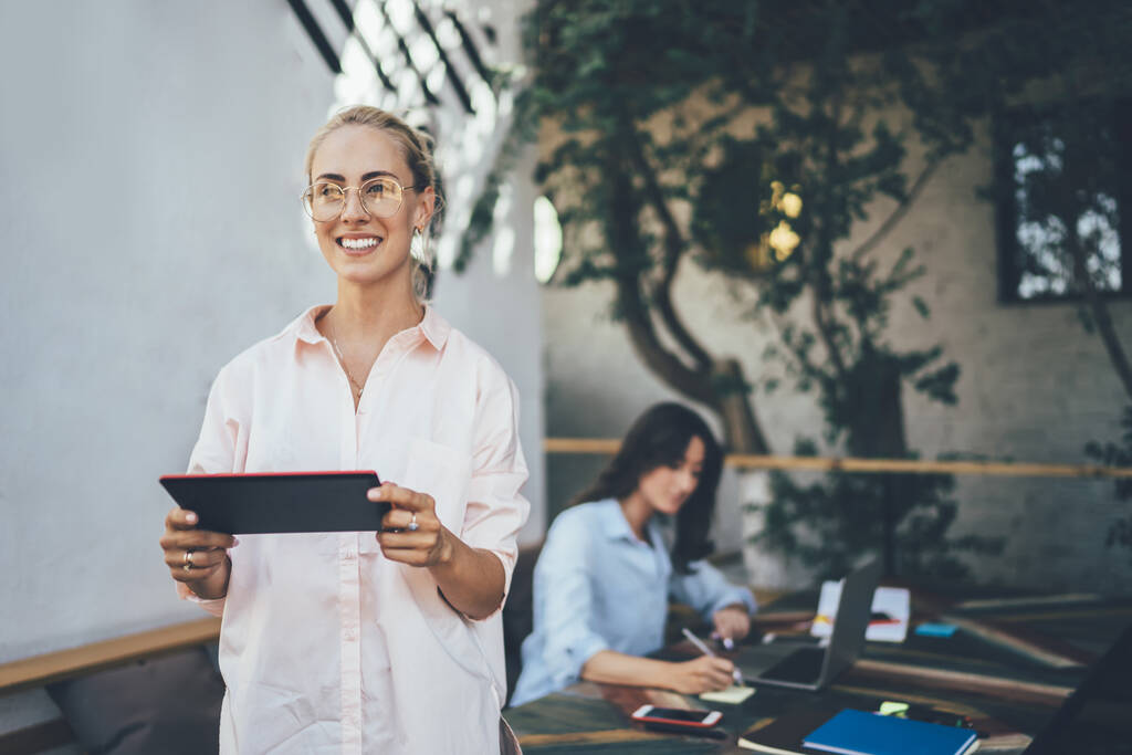 Счастливая женщина с табличкой, улыбающаяся и отводящая взгляд, стоя рядом со столом, и работающая подруга, подрабатывающая на террасе кафе - Фото, изображение