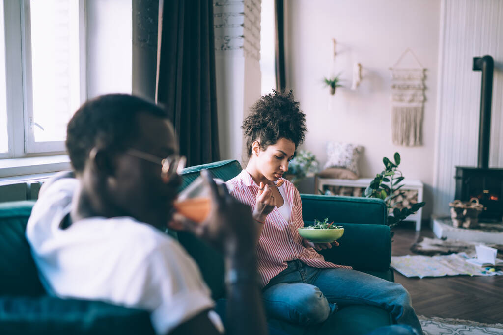 Молода етнічна приваблива жінка в повсякденному одязі і чорний чоловік в окулярах обідають разом, сидячи на зручному дивані в квартирі
 - Фото, зображення