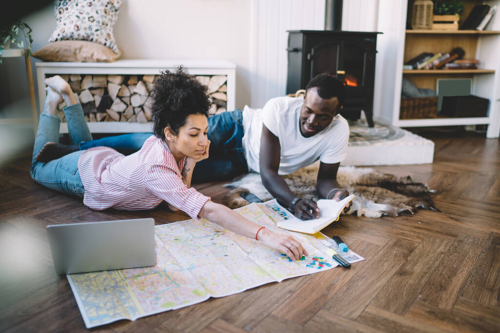 Musta mies ja nainen, jolla on muistikirja ja kartta suunnittelu matka reitti, kun makaa lattialla lähellä kannettavaa kodikasta - Valokuva, kuva