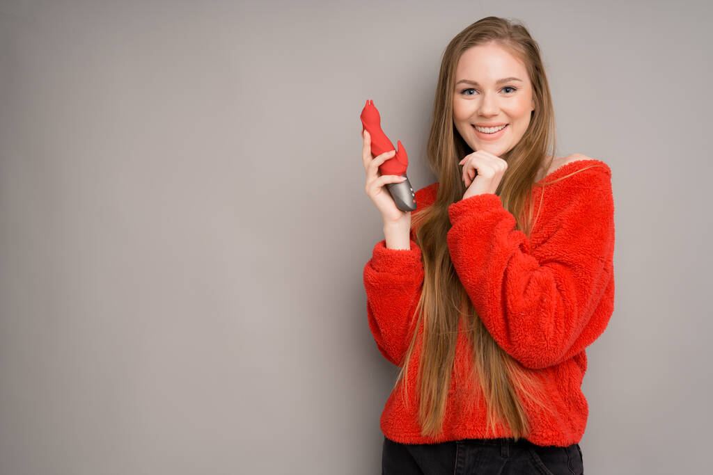An attractive girl of European appearance in a red sweater happily holds a red red dildo in her hands holding her chin with one hand - Φωτογραφία, εικόνα