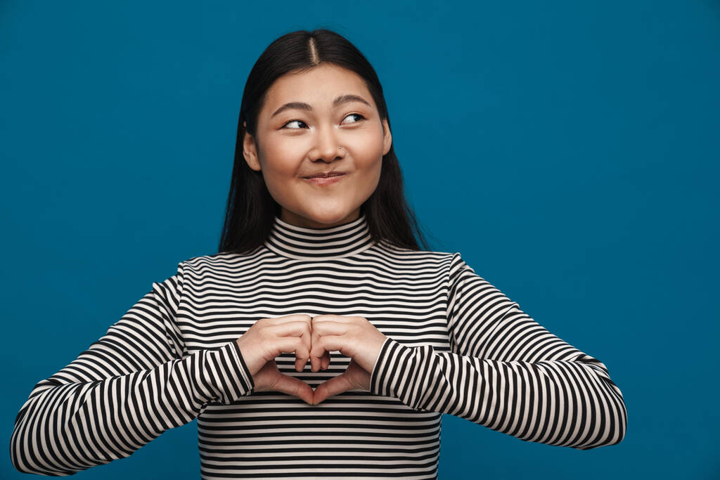 Brunette asian teen girl showing heart gesture at camera isolated over blue background - Photo, Image