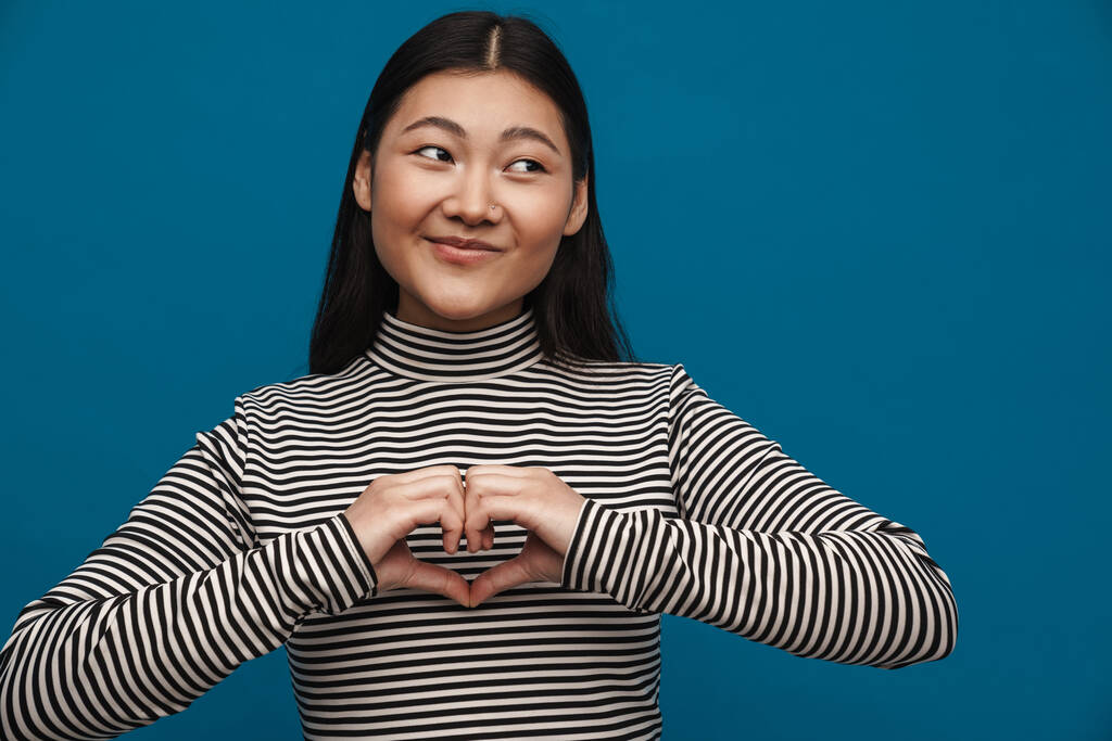 uśmiechnięta azjatycki nastolatek dziewczyna pokazując serce gest w aparat odizolowany na niebieskim tle - Zdjęcie, obraz