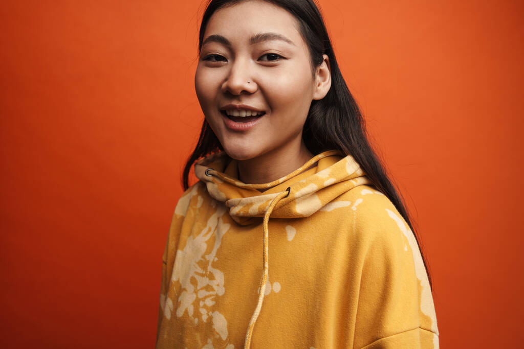 Дівчинка азіатського підлітка, одягнена в капюшон, посміхається і дивиться на камеру, ізольовану над помаранчевим фоном - Фото, зображення