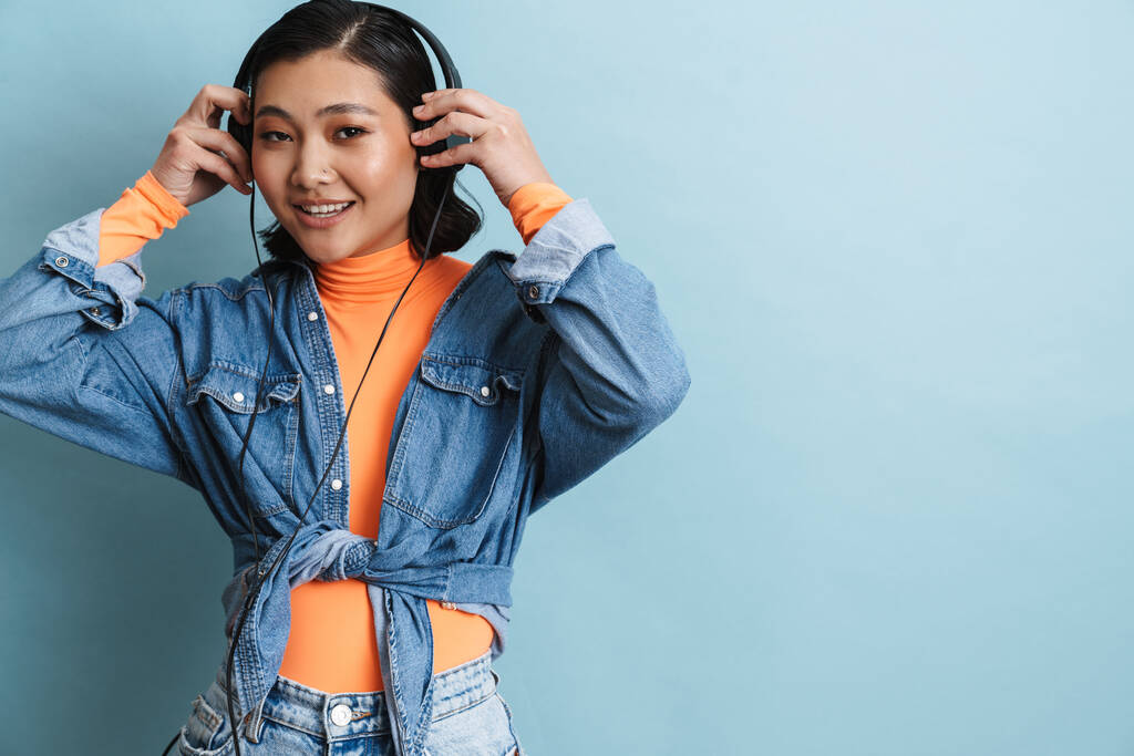 Щаслива усміхнена молода азіатська брюнетка в джинсовій куртці, що стоїть над синім тлом, слухаючи музику з навушниками
 - Фото, зображення