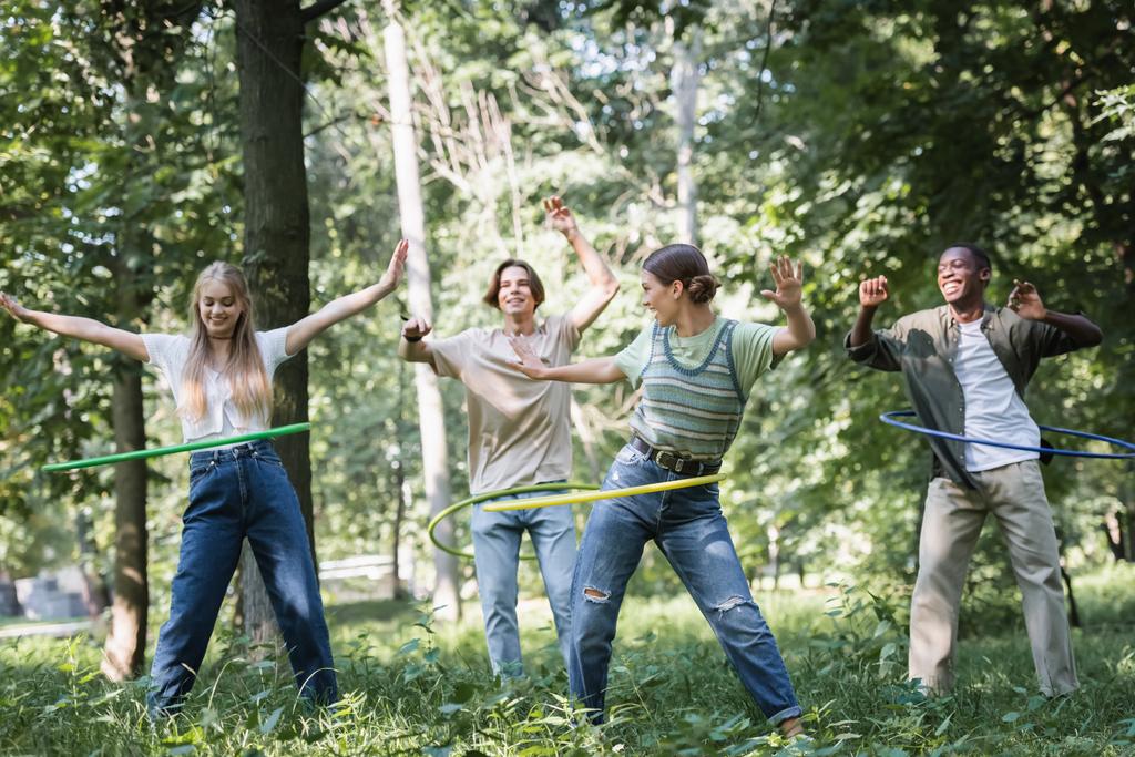 Cheerful multiethnic teenagers twisting hula hoops on grass  - Photo, Image