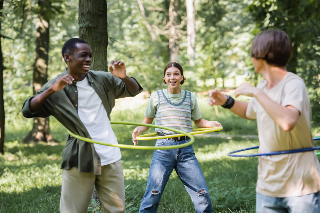 Sonriente chica sosteniendo hula hoop cerca interracial amigos en hierba  - Foto, Imagen