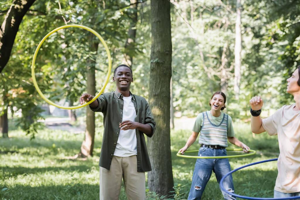 Lächelnder afrikanisch-amerikanischer Teenager mit Hula-Hoop-Reifen bei Freunden im Park  - Foto, Bild