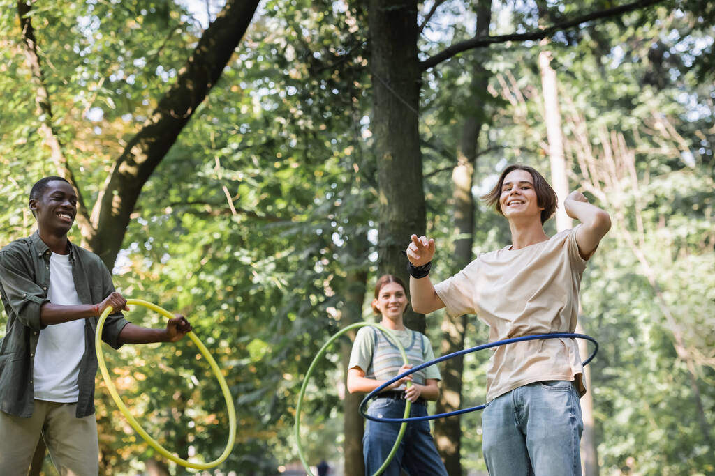 Niski kąt widzenia międzyrasowych nastolatków kręcących hula hoops w parku  - Zdjęcie, obraz