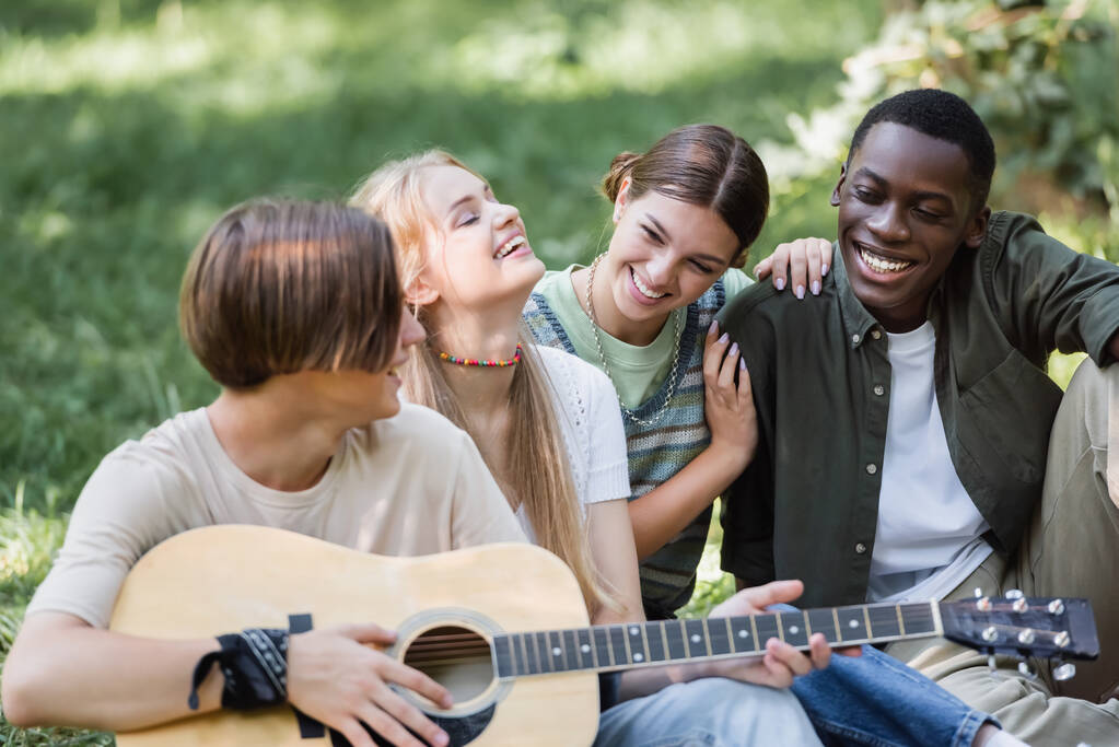 Usmívající se mezirasoví teenageři s akustickou kytarou tráví čas v parku  - Fotografie, Obrázek