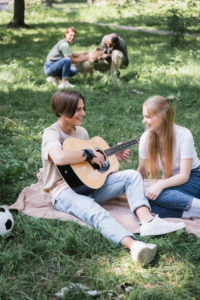 Улыбающиеся подростки, играющие на акустической гитаре рядом с размытыми друзьями с собакой на газоне  - Фото, изображение