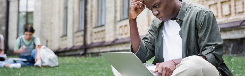 Estudiante africano americano pensativo usando el ordenador portátil al aire libre, bandera  - Foto, Imagen