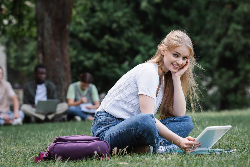 Lächelnder Student hält digitales Tablet in der Hand und blickt auf Rasen in die Kamera  - Foto, Bild