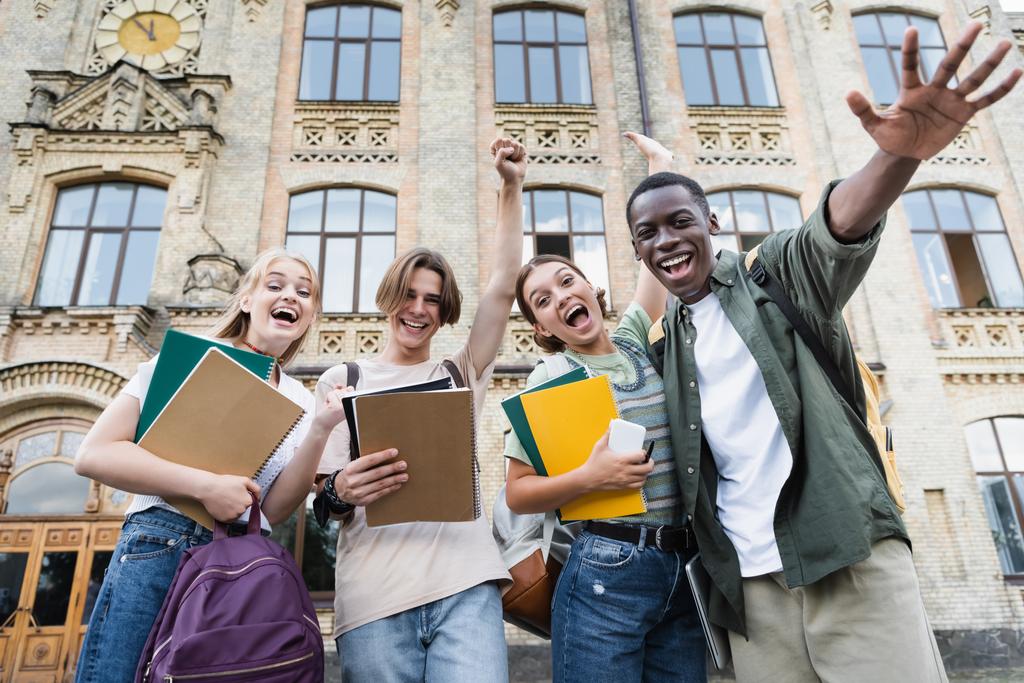 Low angle view van opgewonden interraciale studenten met notebooks op zoek naar camera buiten  - Foto, afbeelding
