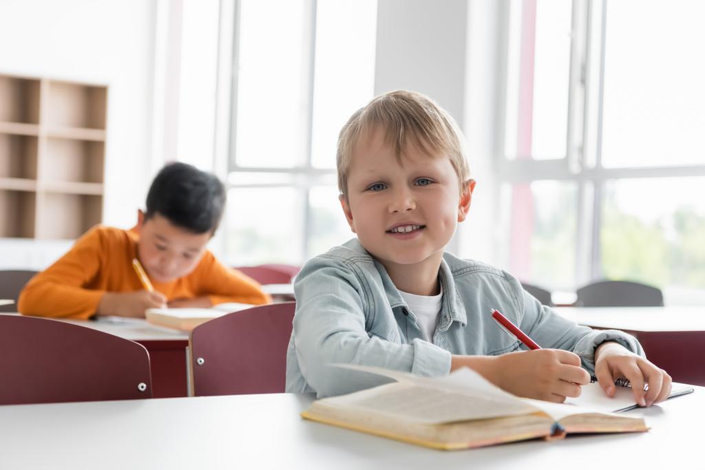 gelukkig schoolkind glimlachen op camera in de buurt aziatische jongen schrijven op wazig achtergrond - Foto, afbeelding