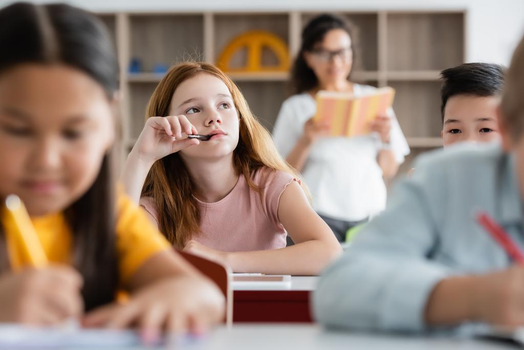 attent roodharige meisje houdt pen in de mond terwijl weg te kijken tijdens de les - Foto, afbeelding
