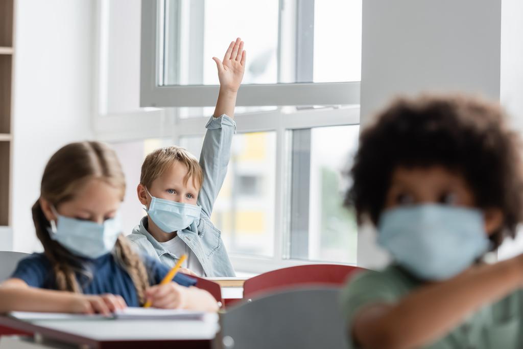 colegial en máscara médica levantando la mano cerca borrosa compañeros de clase multiétnicos durante la lección - Foto, Imagen
